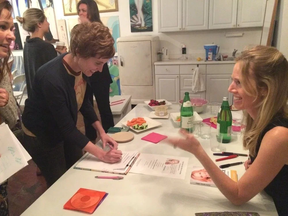 Group of women gathered around a kitchen counter, with two women having a conversation, one signing a book.