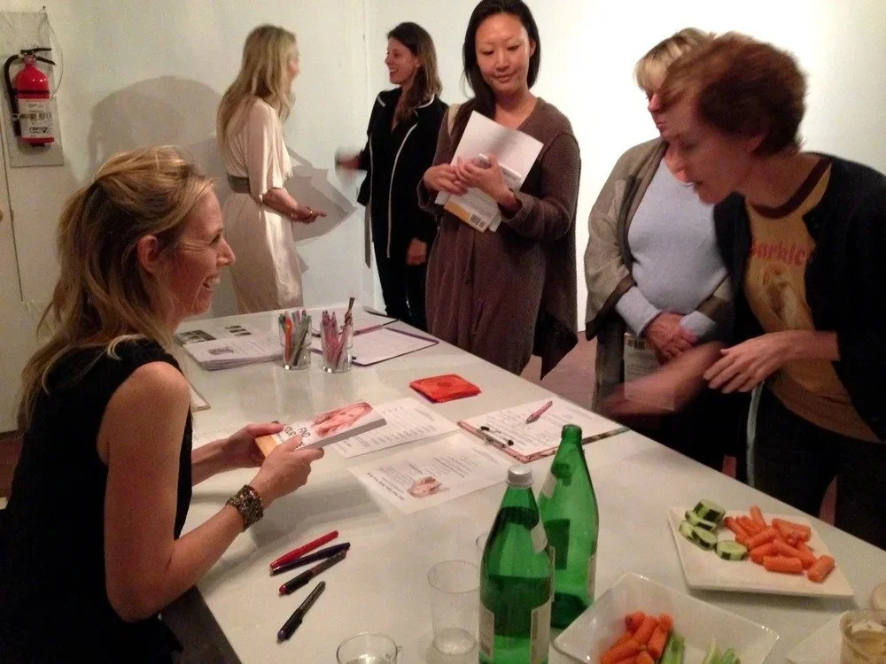 A woman sitting at a table signing a book for a group of women standing around her in a room with white walls. The table has water bottles, papers, and snacks including sliced cucumbers and carrots.