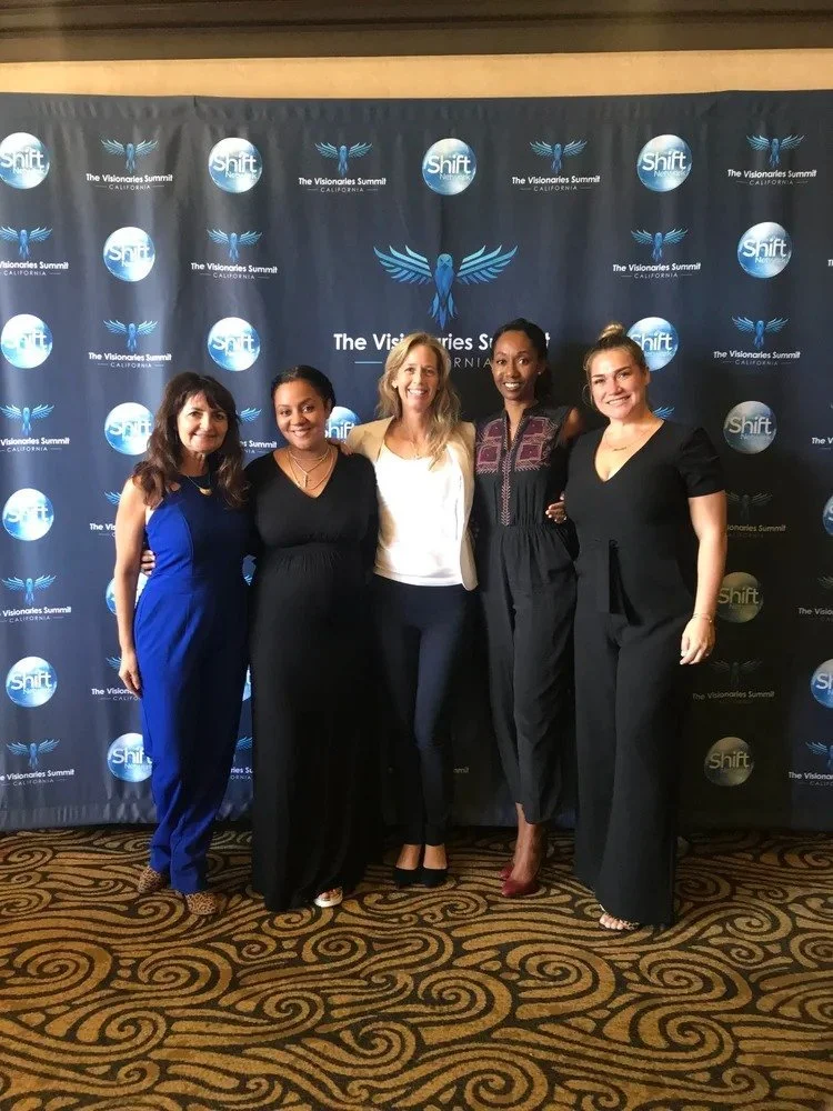 Five women standing together in front of a backdrop with blue logos and text that reads 'The Visionaries Summit' and 'Shift'. They are smiling, dressed in formal attire, with the woman in the middle wearing a white blazer, and others in black or blue