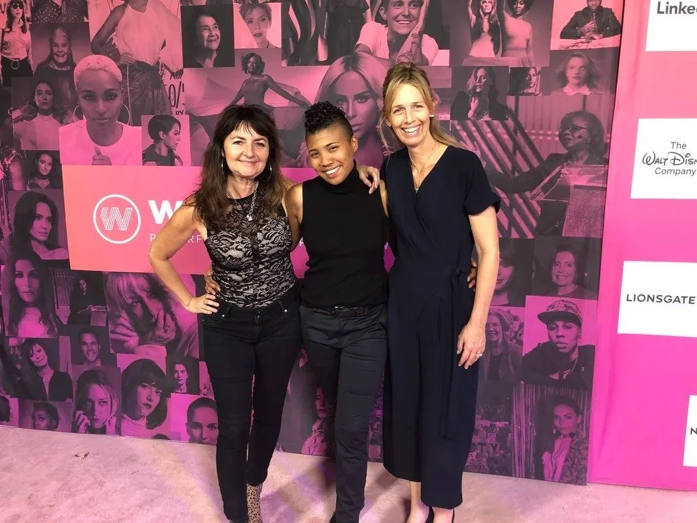 Three women standing together, smiling, at an event with a pink and black collage backdrop featuring images of women.