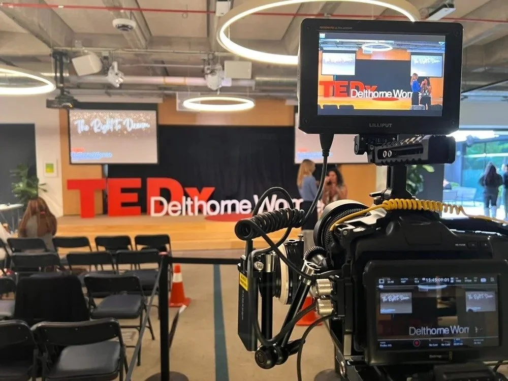 An empty auditorium with a stage at TEDxDelthomeWor event. There is a large red TEDx sign and a screen on the stage displaying the same. A film camera is set up in the foreground capturing the event.