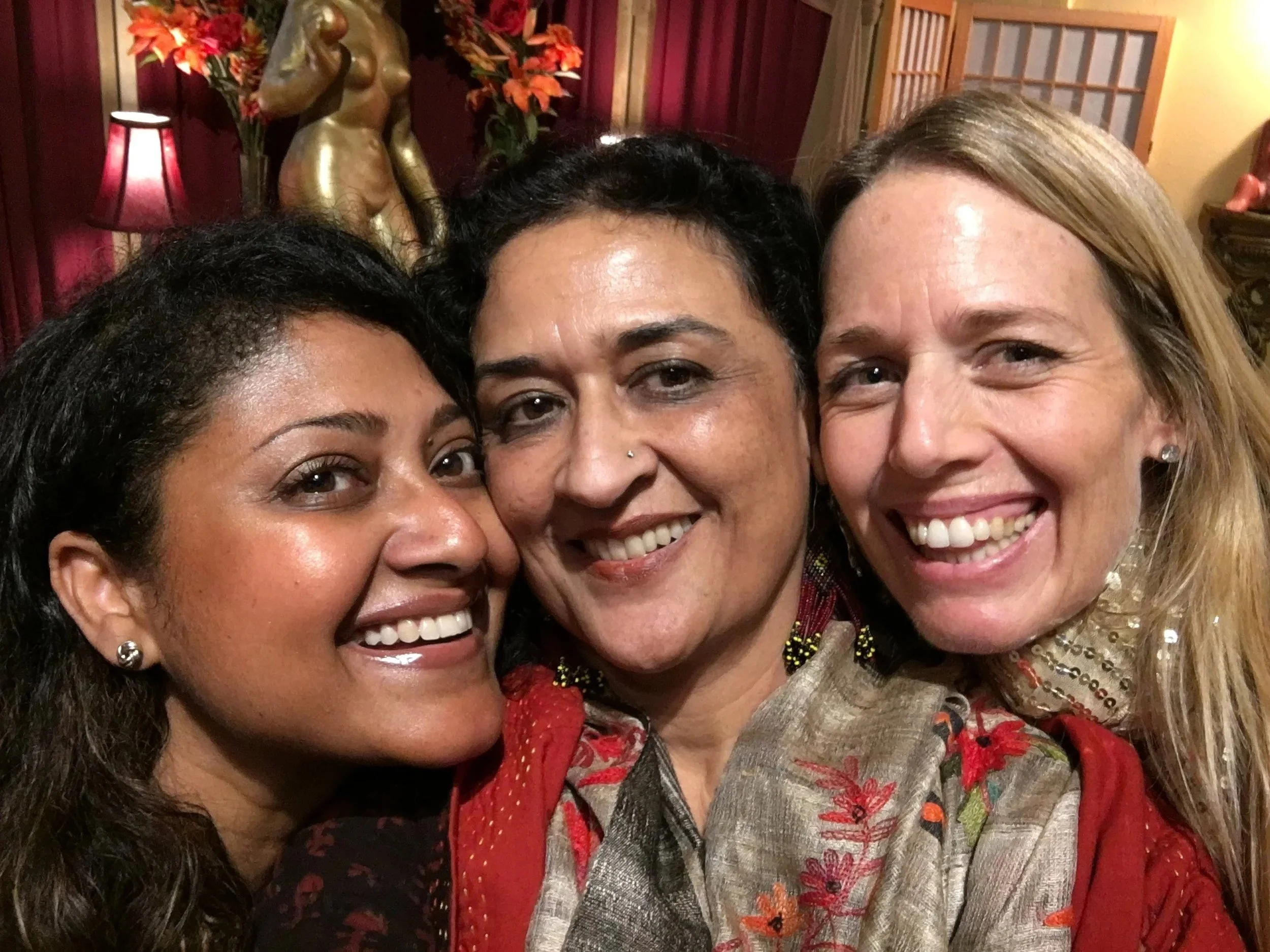 Three women smiling closely together for a selfie in an ornately decorated room with flowers, lamps, and a sculpture in the background.