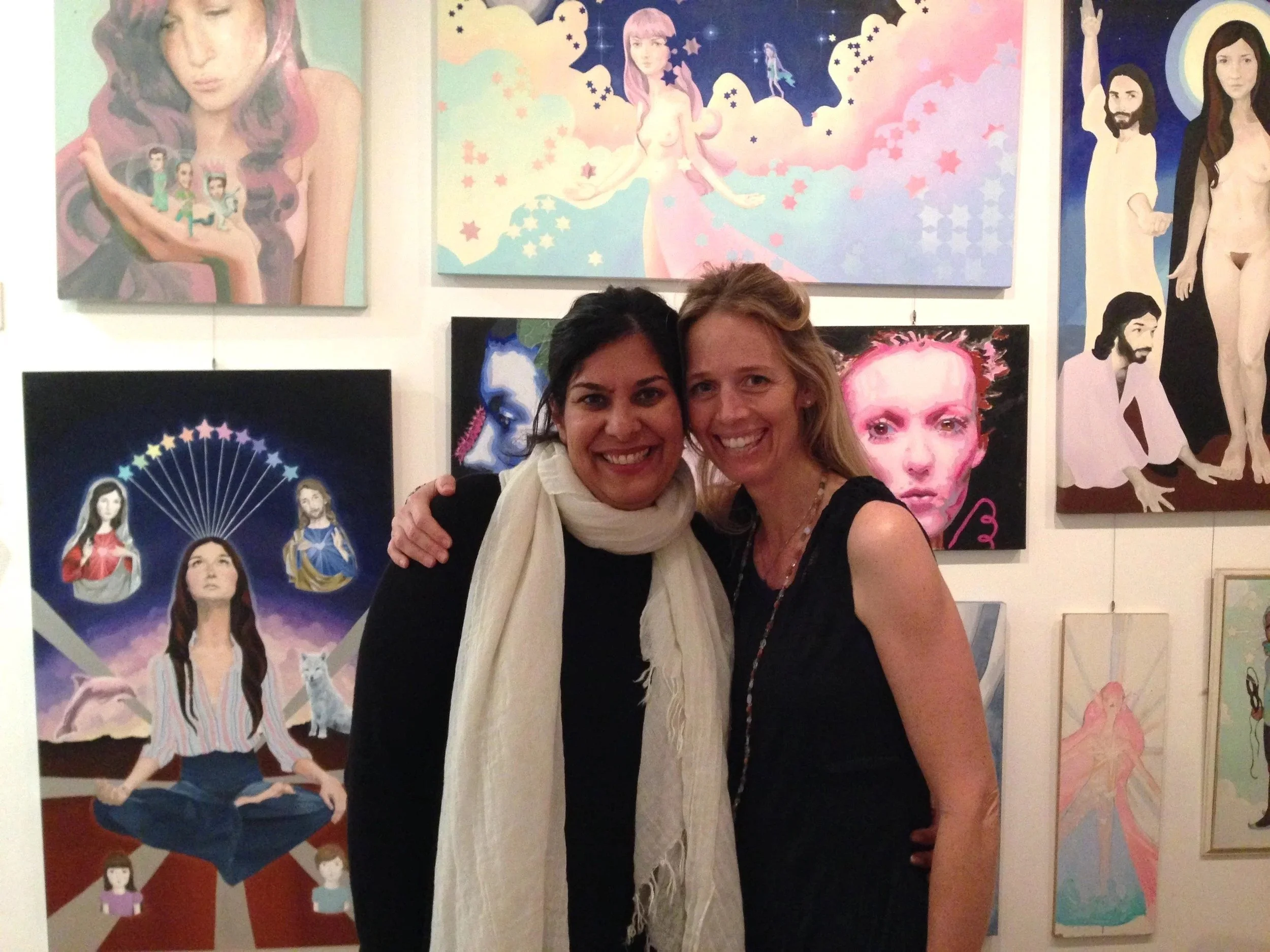 Two women smiling and hugging at an art gallery with colorful paintings on the wall behind them.
