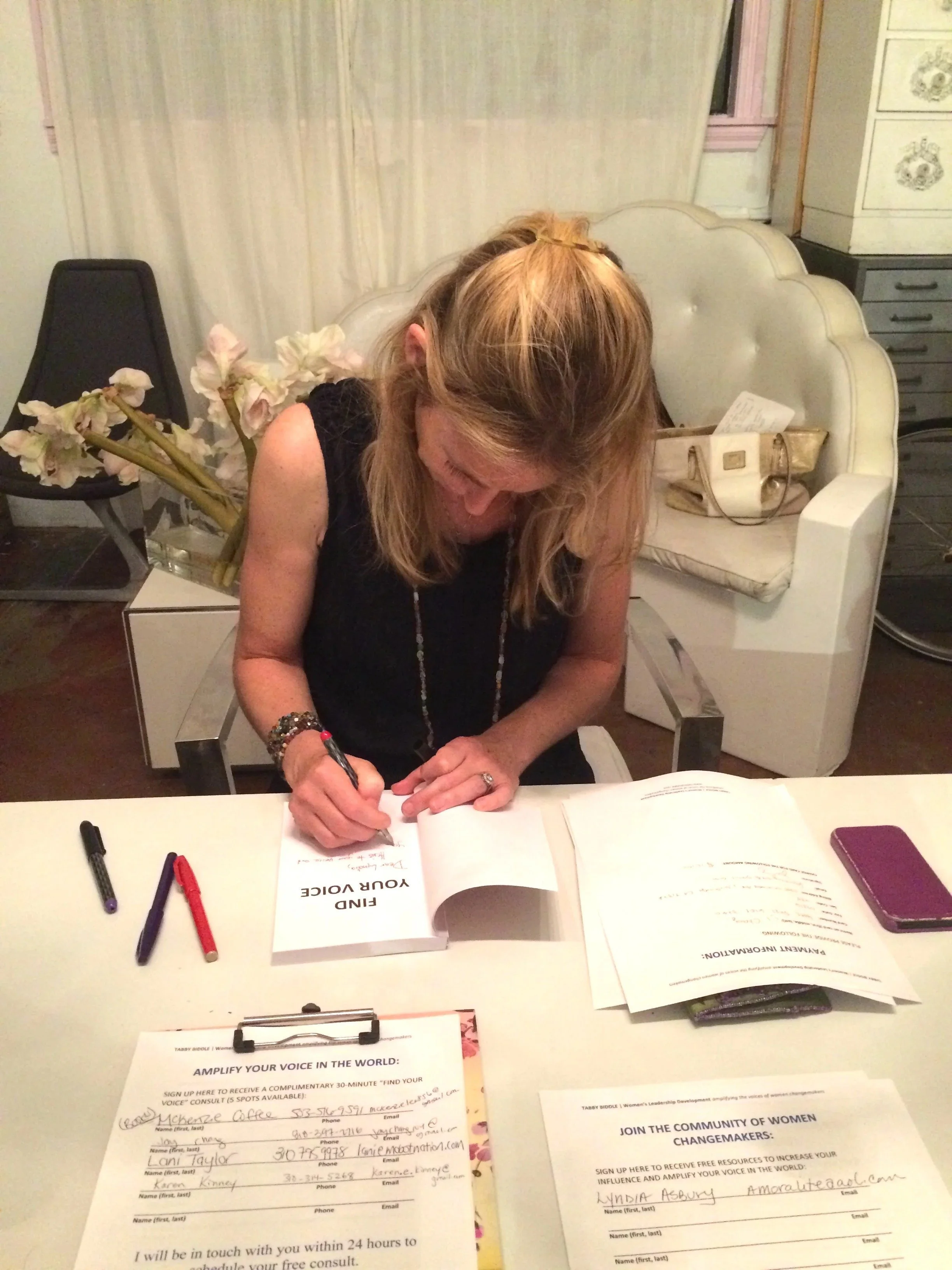 A woman with blonde hair signing a book titled 'Find Your Voice' at a table with papers, markers, and a phone, in a room with a white couch, flowers, and chairs.