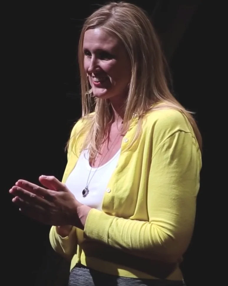 Woman with long blonde hair smiling, wearing a yellow cardigan and a white shirt, standing against a dark background.