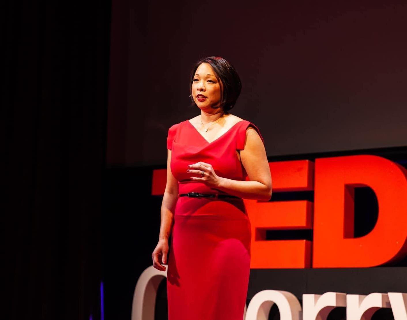 A woman in a red dress giving a presentation on stage with a large red TED logo in the background.