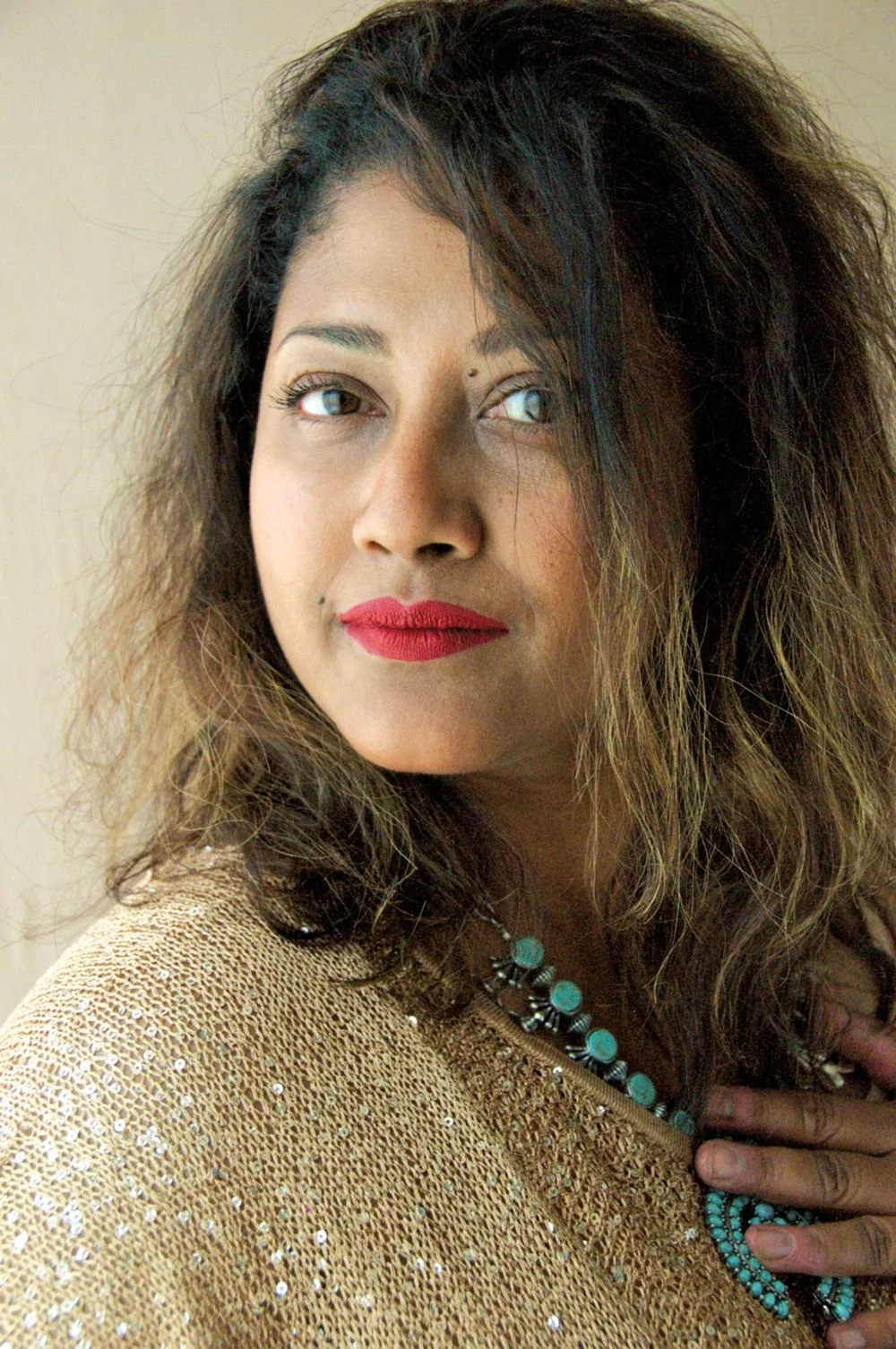 A woman with voluminous, wavy hair, wearing red lipstick, a gold sequined top, and turquoise jewelry, looking slightly to the side.