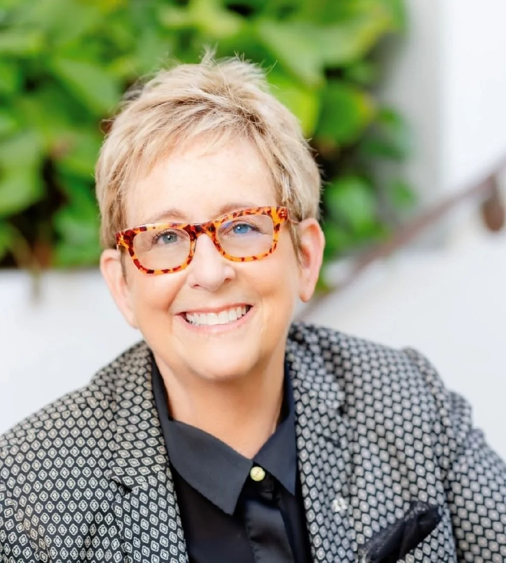 Smiling woman with short blonde hair, tortoiseshell glasses, black button-up shirt, patterned blazer, and a background of green foliage.