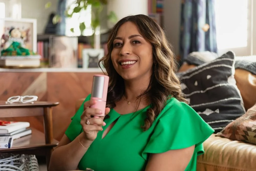 A woman with wavy brown hair in a green top smiling and holding a pink microphone or recording device in a living room setting.