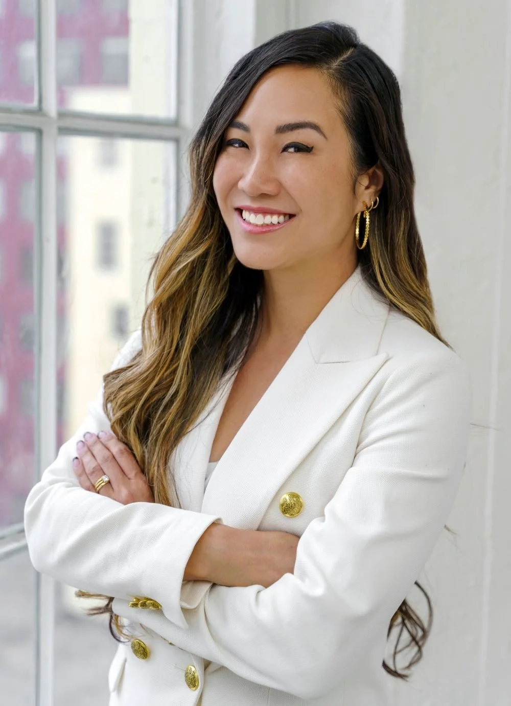 A woman with long wavy hair, smiling, wearing a white blazer with gold buttons, gold hoop earrings, and a ring on her left hand, standing near a window.
