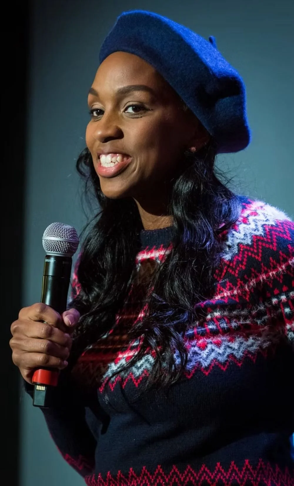 A woman with dark curly hair wearing a blue beret and a red, white, and blue patterned sweater, holding a microphone and smiling.