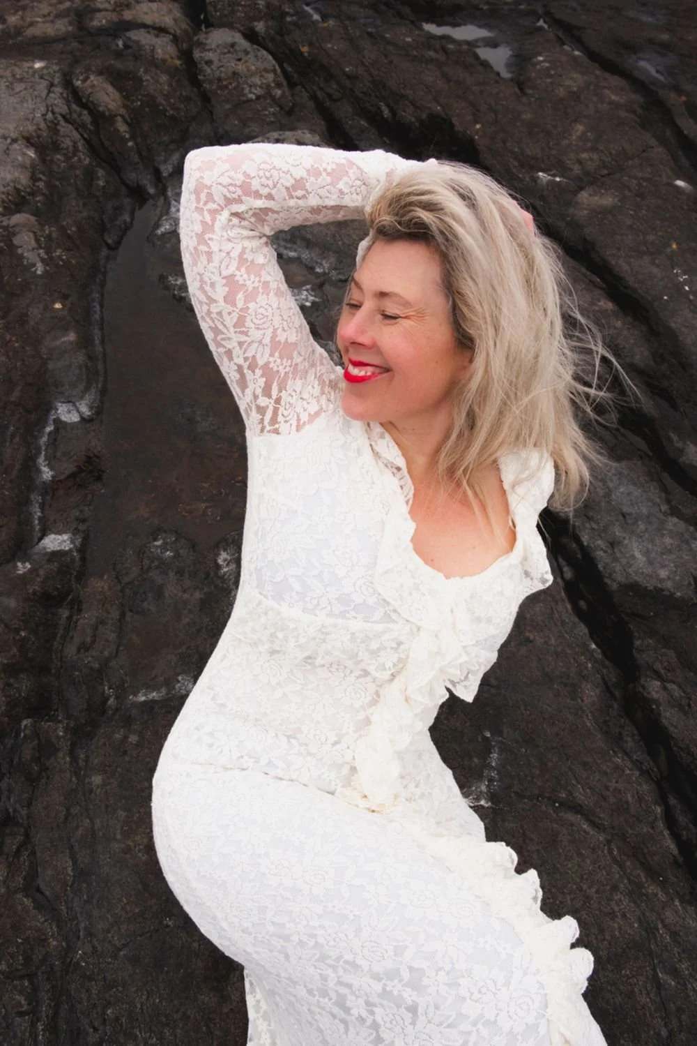A woman with blonde hair wearing a white lace dress, smiling with eyes closed, standing against dark rocks.