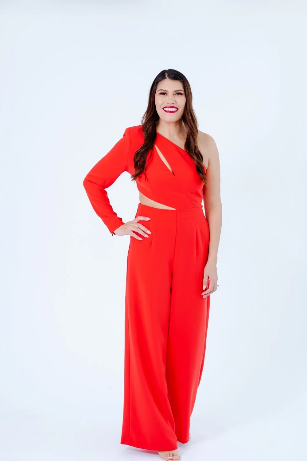 Woman in a red stylish jumpsuit with one shoulder cutout, standing against a plain white background, smiling with long dark hair
