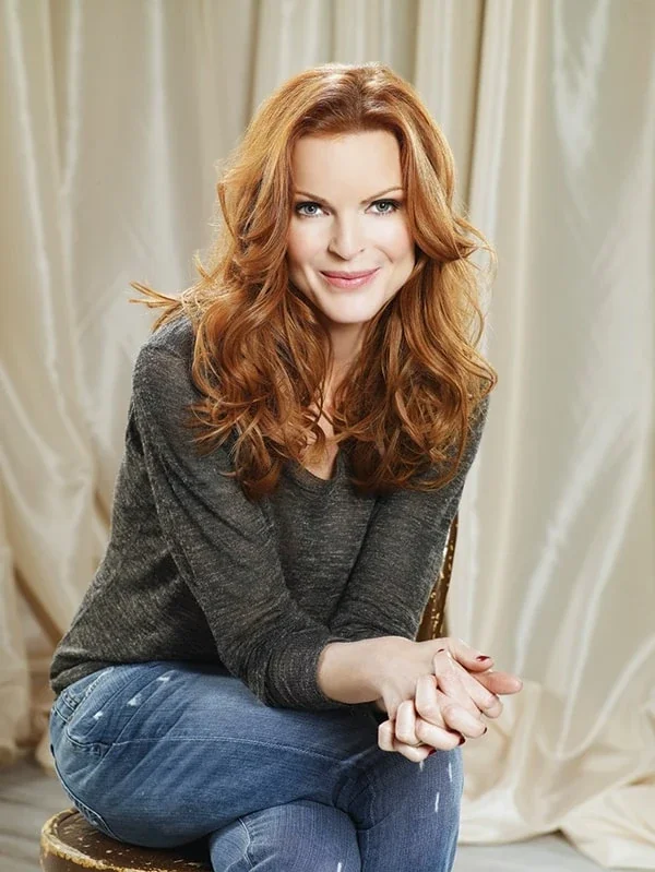 A woman with long wavy hair, wearing a denim jacket over a patterned dress, sitting on a gray couch in a room with a framed picture on the wall behind her, smiling at the camera.