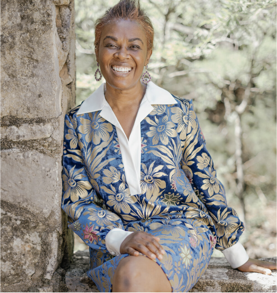 A woman with short hair and earrings, smiling and sitting outdoors near a stone wall surrounded by trees, wearing a blue floral-patterned blazer and skirt with a white shirt.