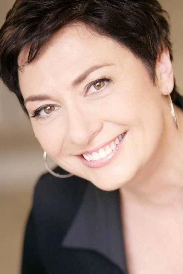 Close-up of a smiling woman with short dark hair, wearing hoop earrings and a black blazer.