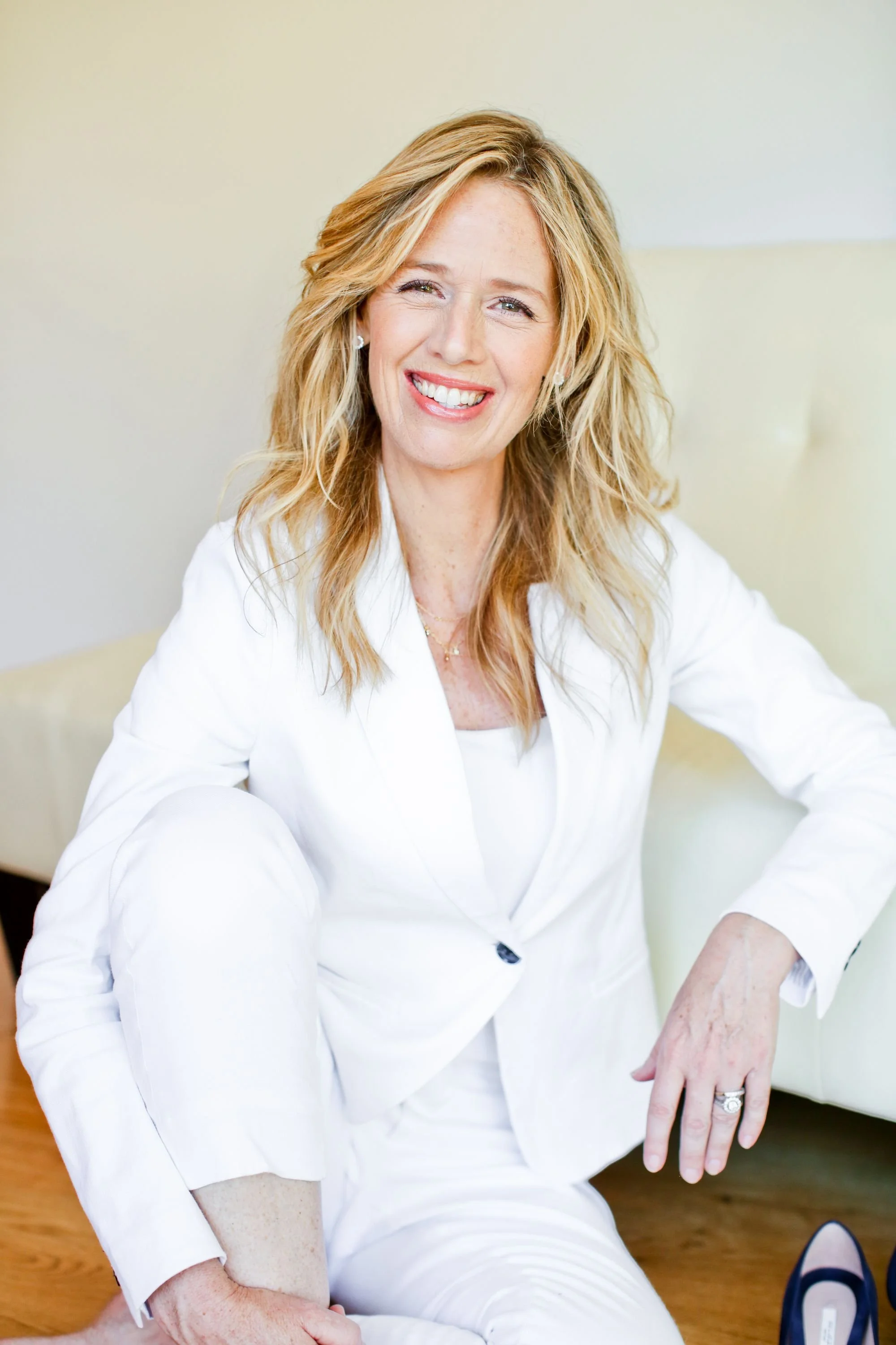 A woman with blonde wavy hair wearing a white suit, smiling and sitting on the floor.