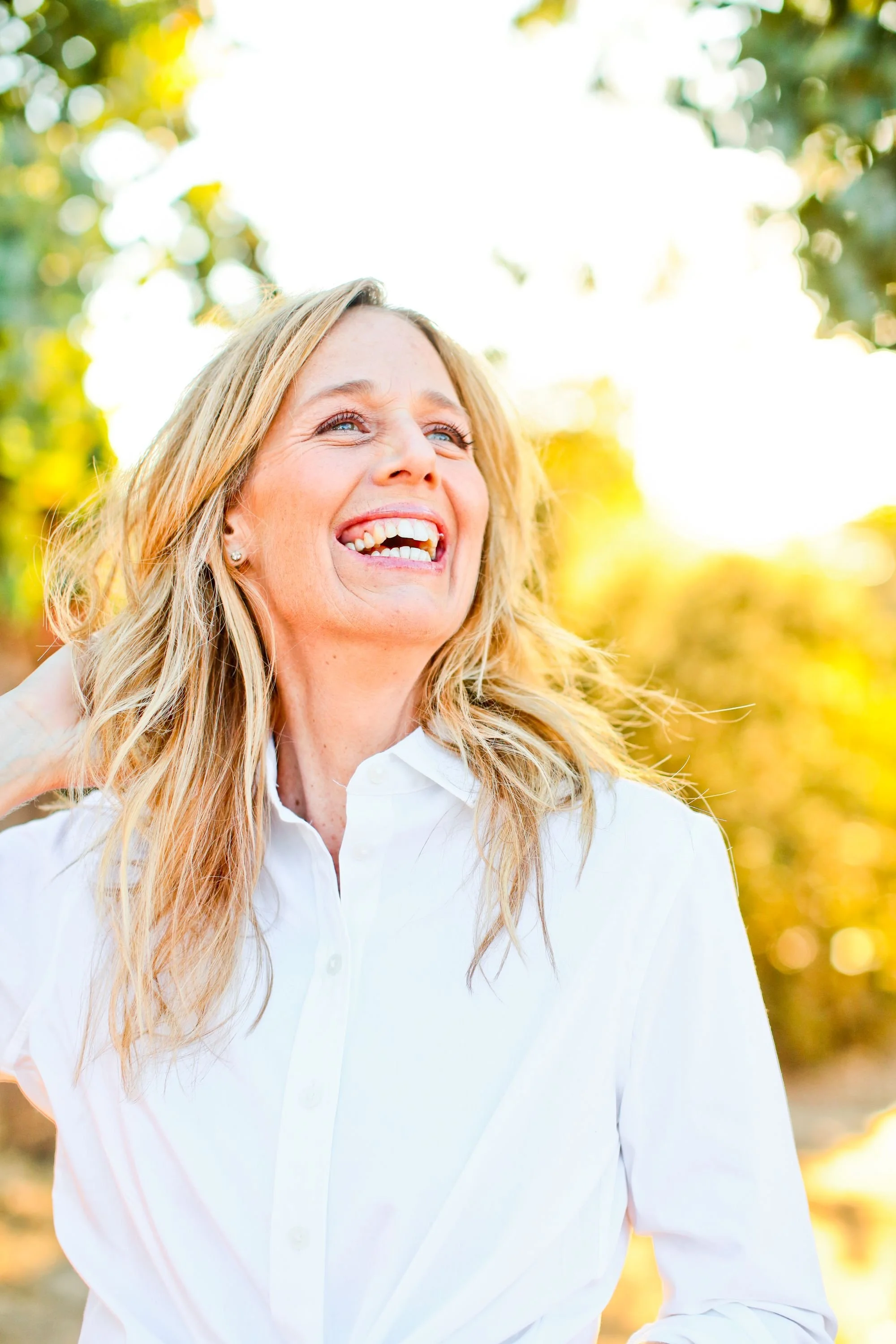 A woman with blonde hair wearing a white shirt, smiling outdoors in bright sunlight with a background of green trees.