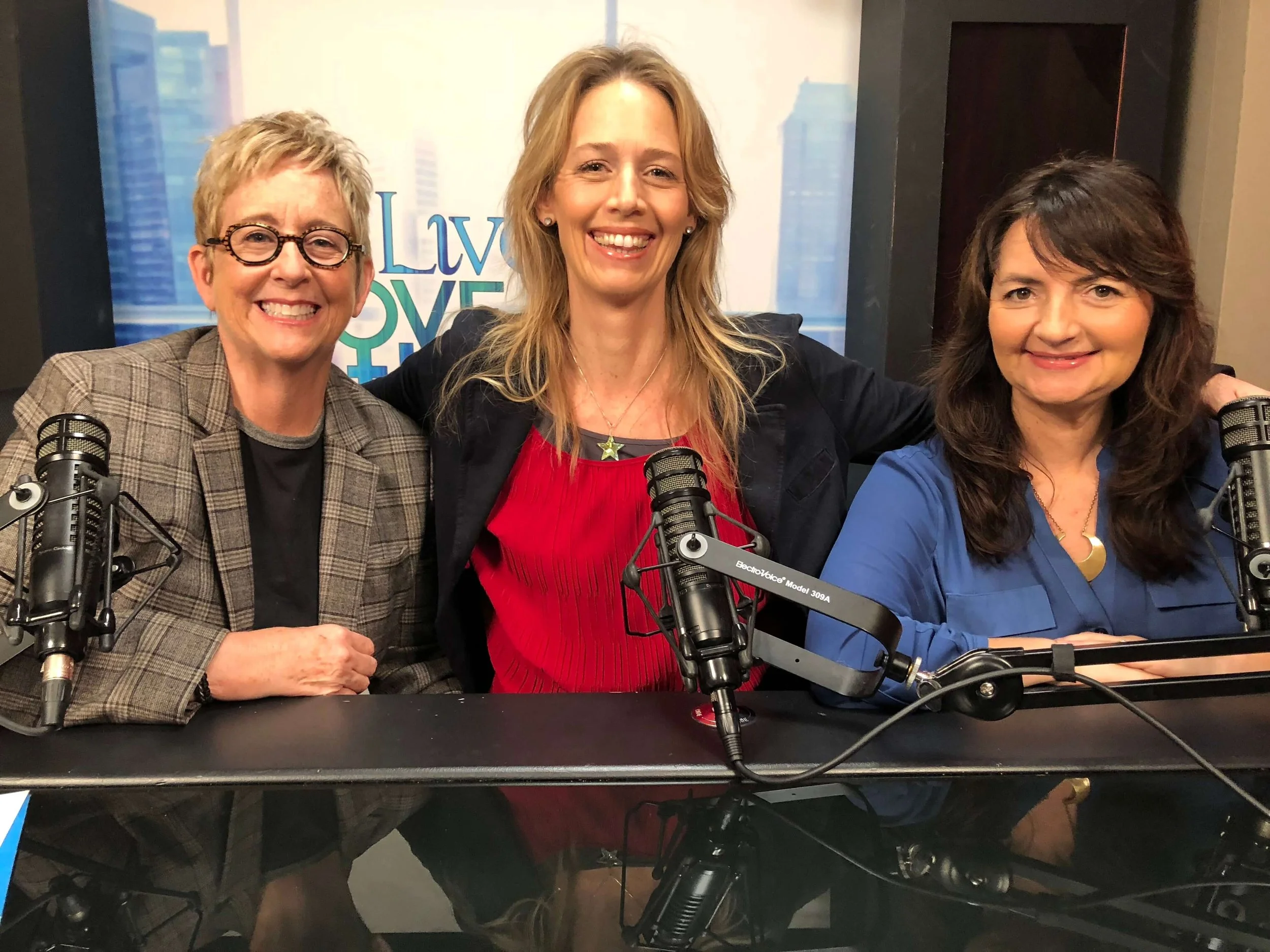 Three women sitting side by side at a table with microphones, smiling for a photo in a radio studio, with a backdrop that reads "Love".