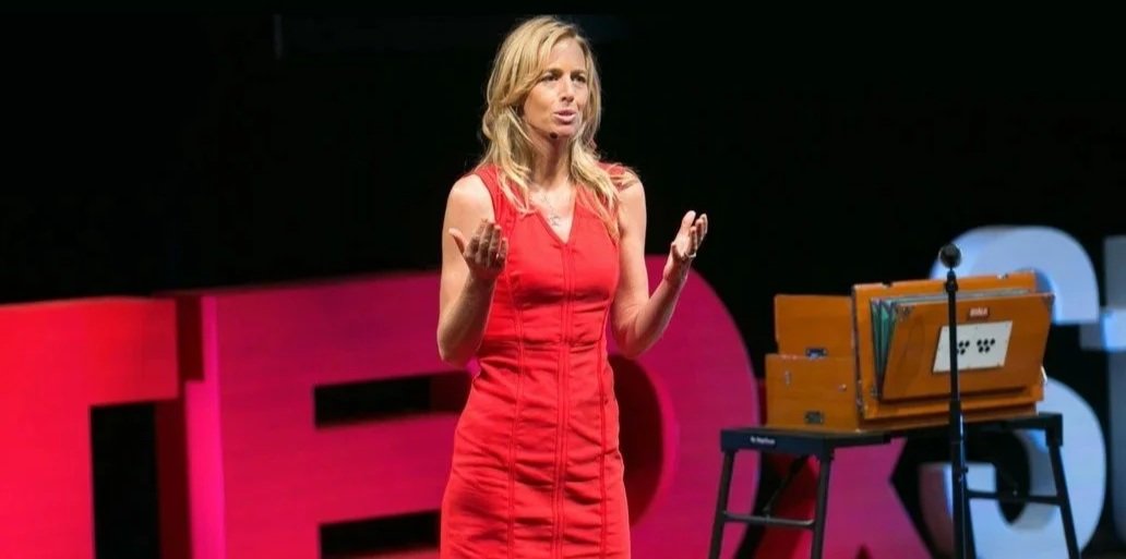 A woman in a red dress giving a TED Talk on stage, with TED letters and a wooden music box on a table beside her.