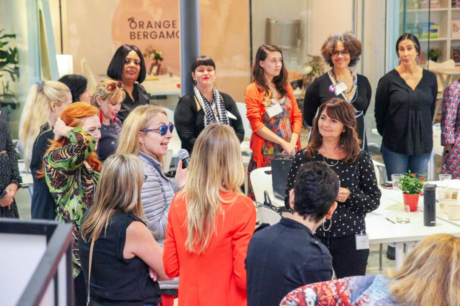 A group of women gathered at an indoor event, one woman with long blonde hair and sunglasses holding a microphone speaking to the group. The women are standing and sitting around a table with drinks and plants, engaging in a discussion or presentation.