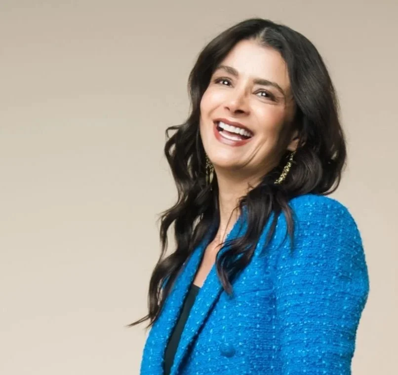 A woman with long dark wavy hair smiling and wearing a blue blazer.