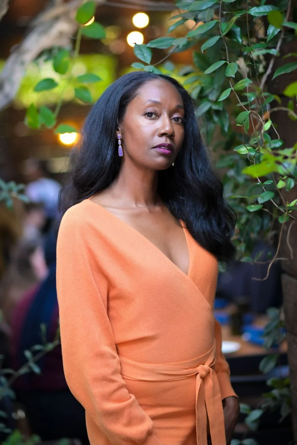 A woman with long black hair and purple earrings wearing an orange wrap dress standing among green plants. Blurred lighting and people in the background.
