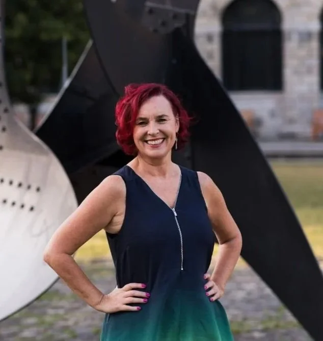 A woman with short, curly red hair smiling and standing outdoors in front of abstract metal sculptures.