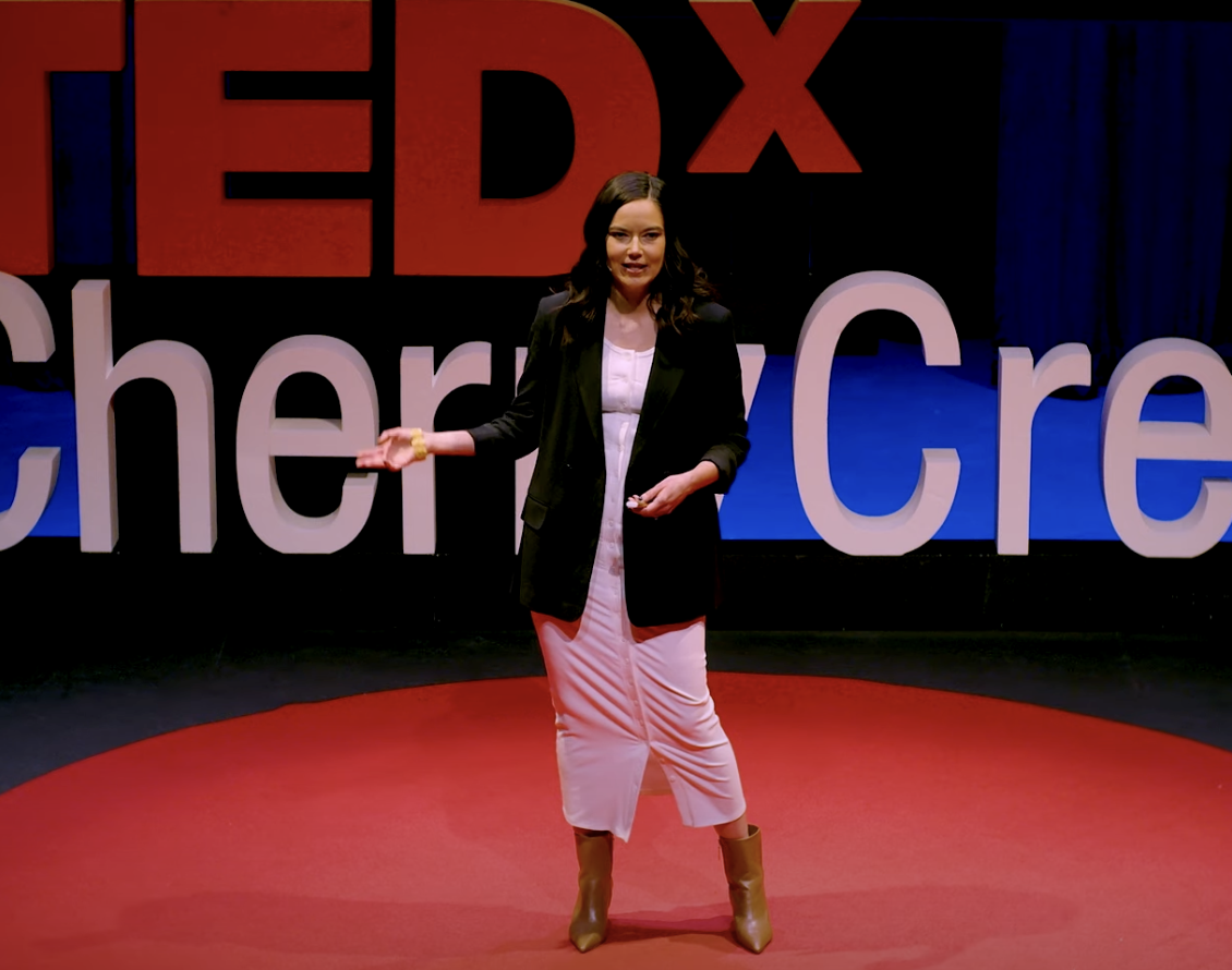A woman in a red dress giving a presentation on a stage with a large TED logo in the background.