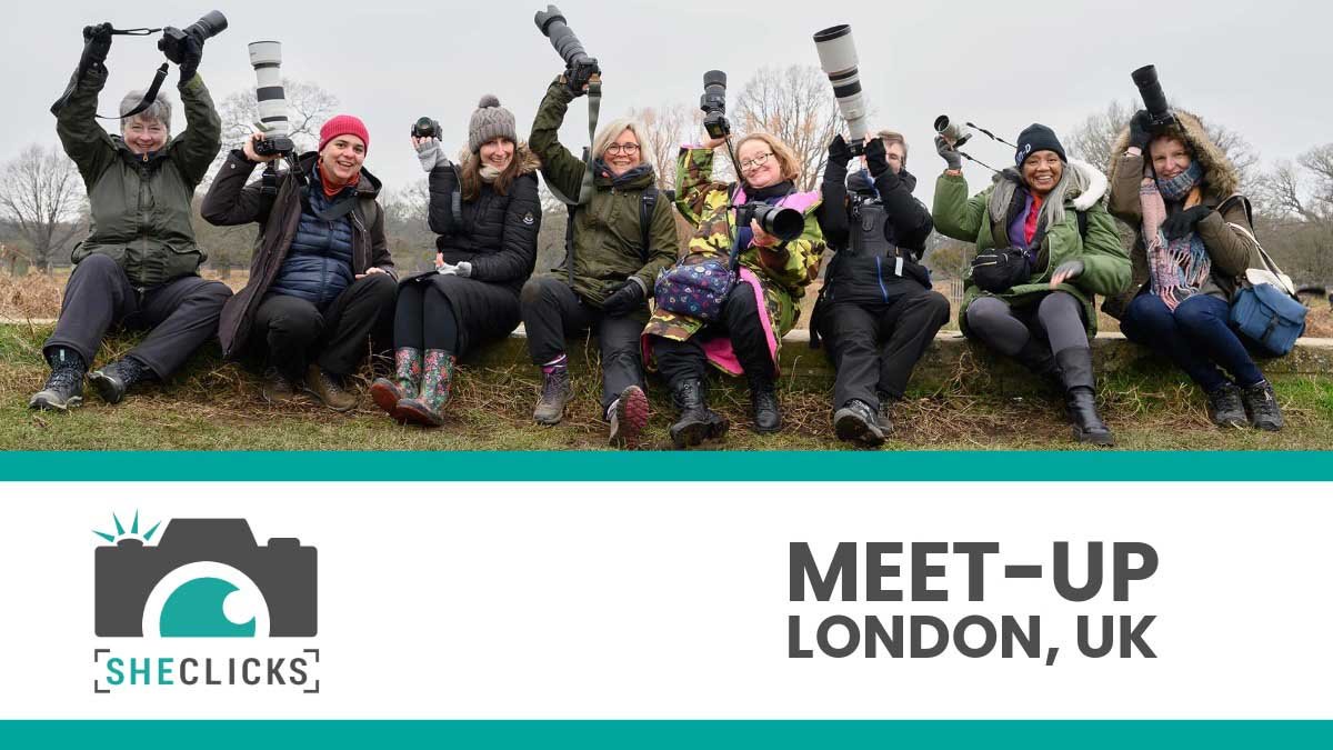 Group of photographers sitting outdoors, smiling and holding up cameras, with text “SheClicks Meet-Up London, UK.”
