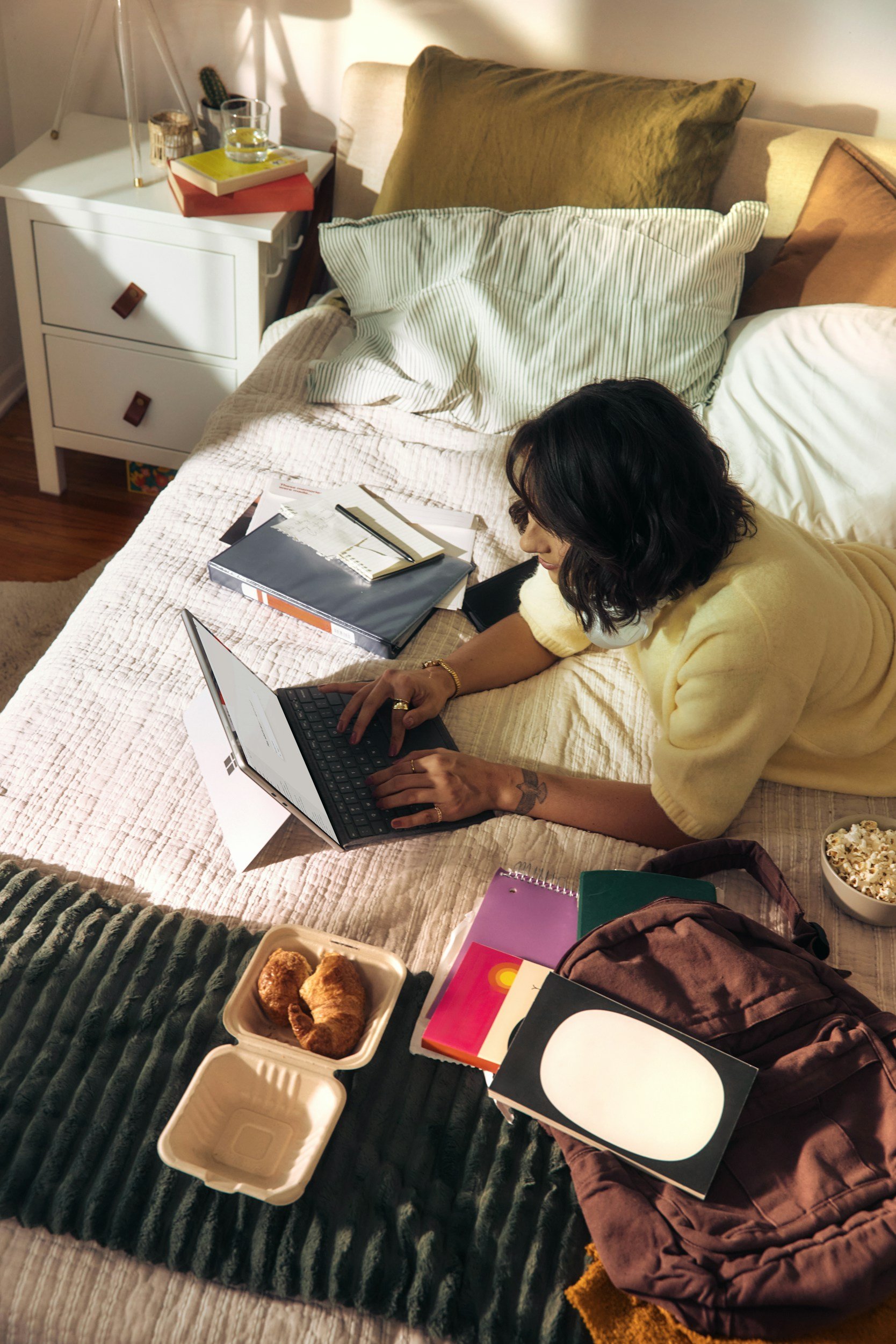 Woman in yellow sweater on her laptop in bed surrounded by snacks