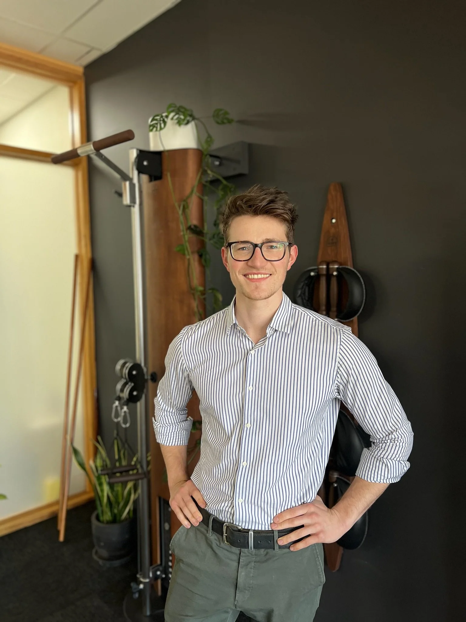 A smiling man in glasses and a striped button-up shirt standing indoors with hands on hips, in front of a dark wall, with exercise equipment and plants in the background.