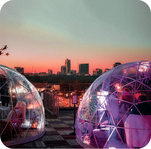 People inside transparent dome tents on a rooftop at sunset with city skyline in the background.