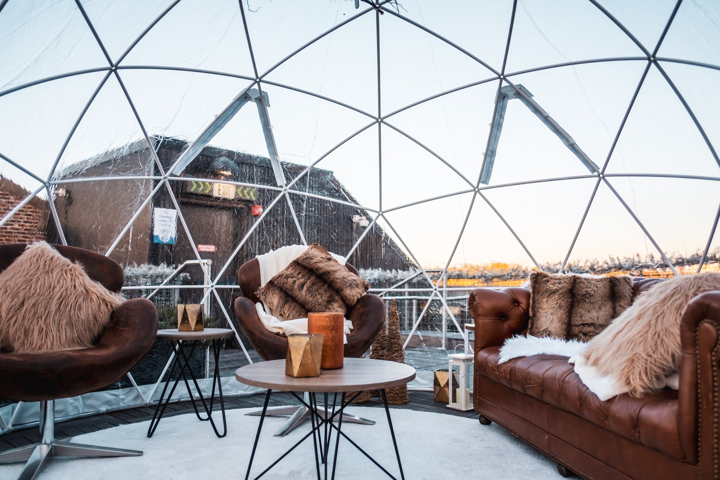 Cozy interior with leather armchairs and sofa with fur pillows, inside a geodesic dome, overlooking water and cityscape at sunset.