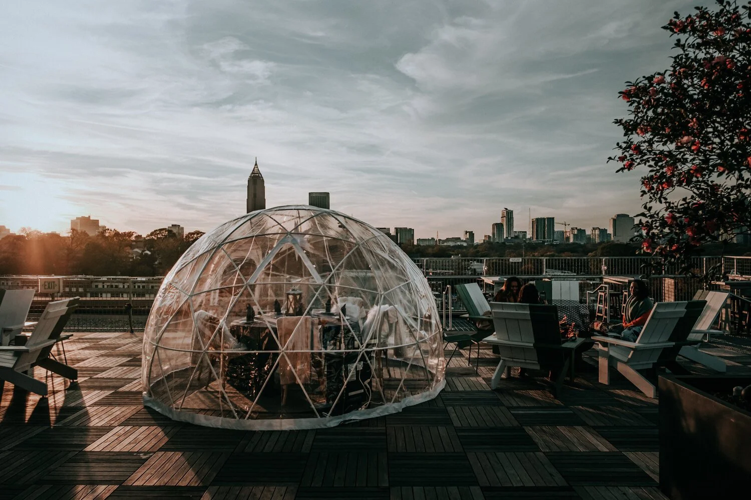 People sitting on chairs and tables outdoors on a rooftop with a city skyline in the background during sunset. There is a clear geodesic dome structure on the wooden deck, and a tree with pink flowers on the right side.