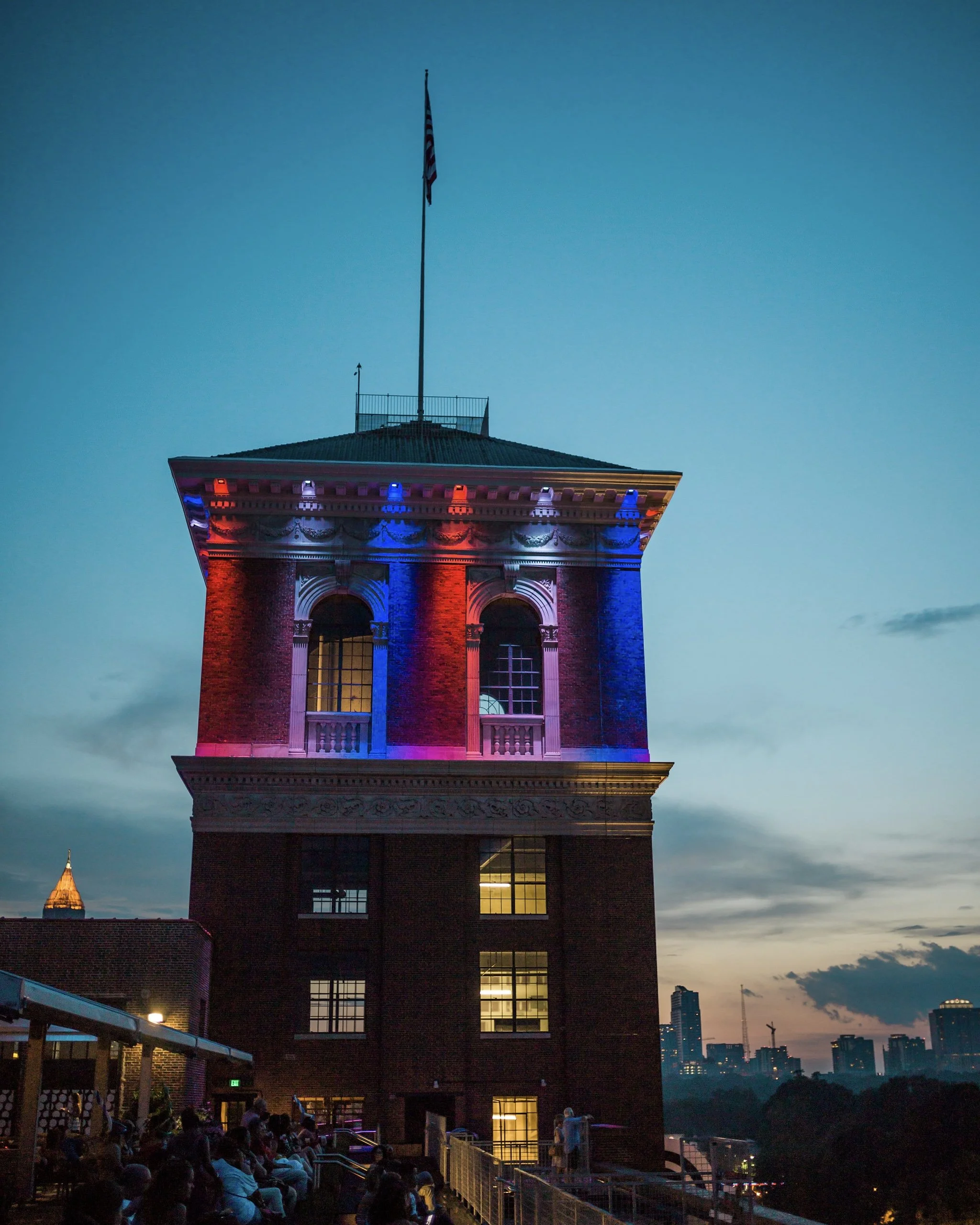 The image shows a tall building with a triangular top, illuminated with red, blue, and purple lights. An American flag is flying from the top of the building, and the sky in the background is transitioning from sunset to dusk with dark clouds and city skyline in the distance.