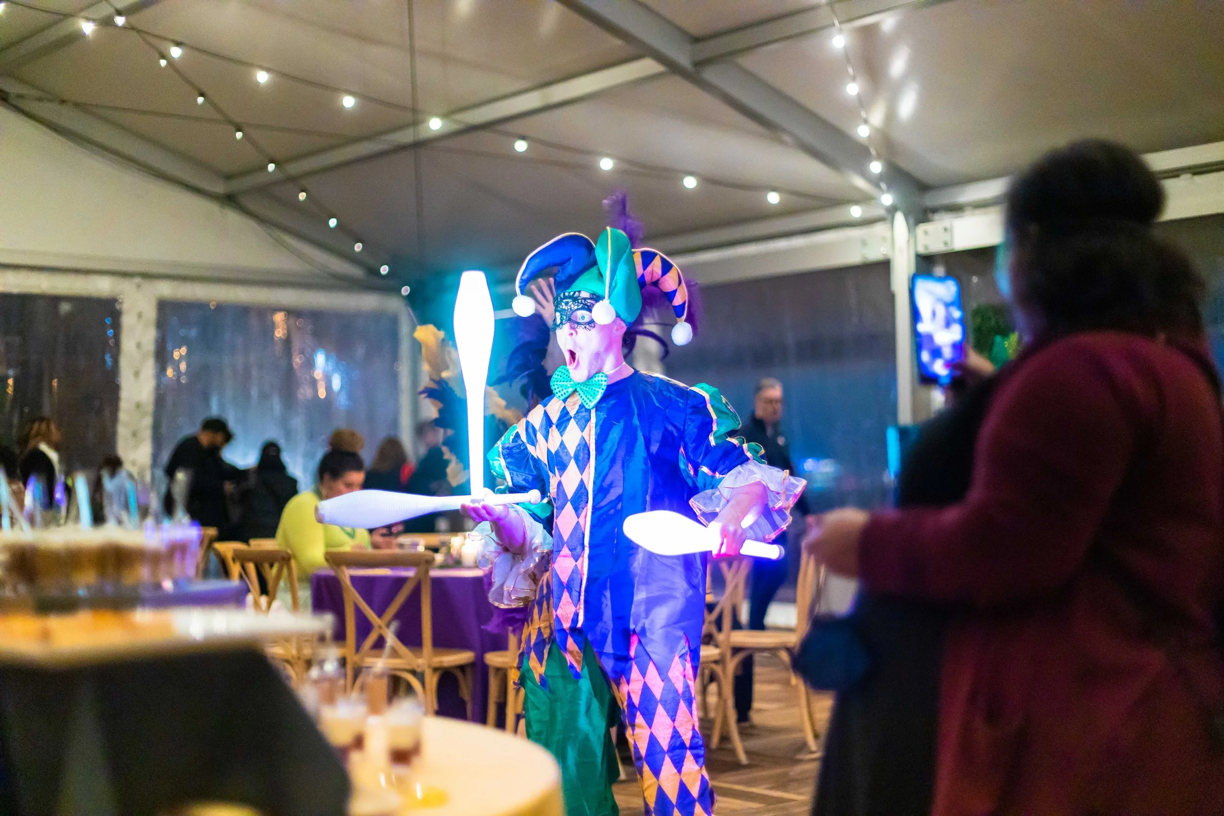 Person dressed as a jester with a colorful costume and hat, juggling glowing clubs at a party or event inside a tent with string lights, with people in the background and a woman taking a photo.