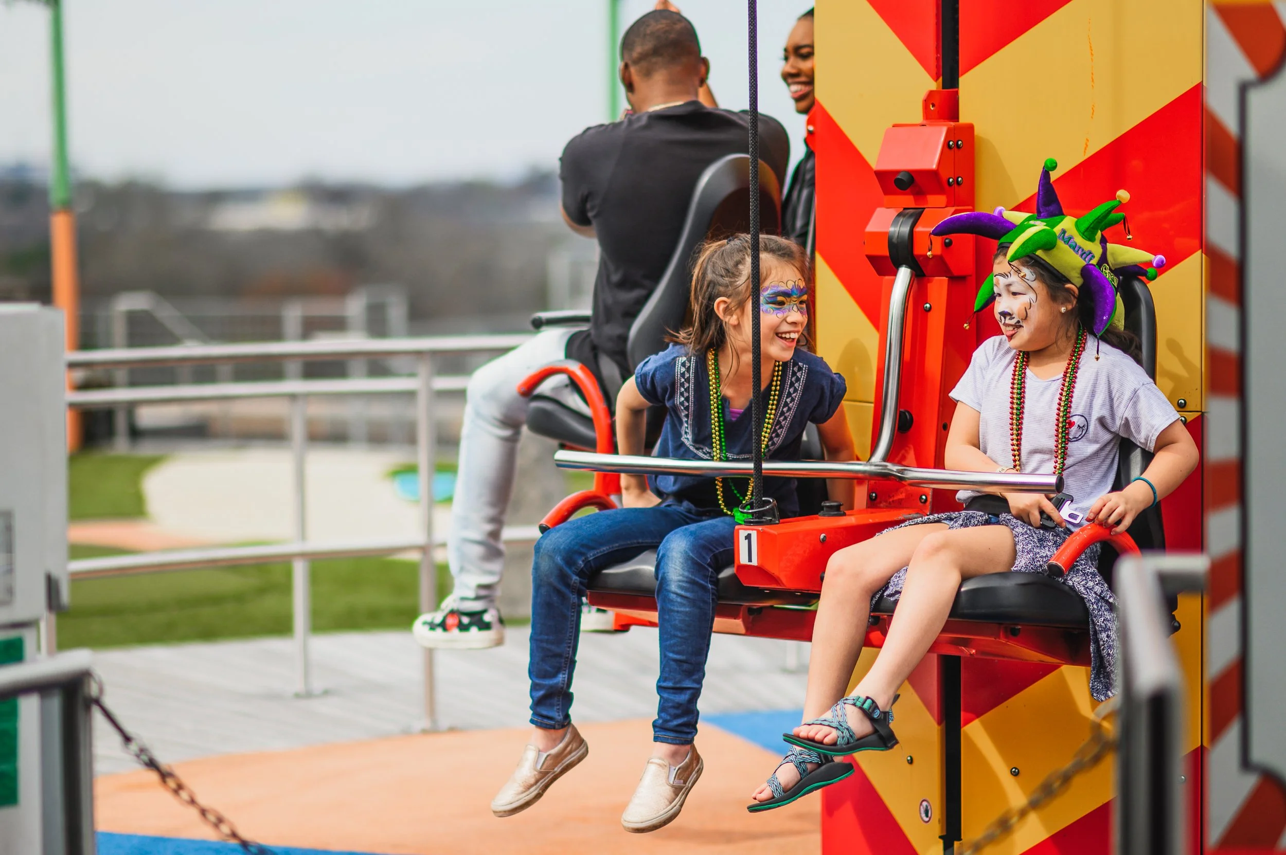 Children with face paint and colorful hats riding a carnival ride at an amusement park.