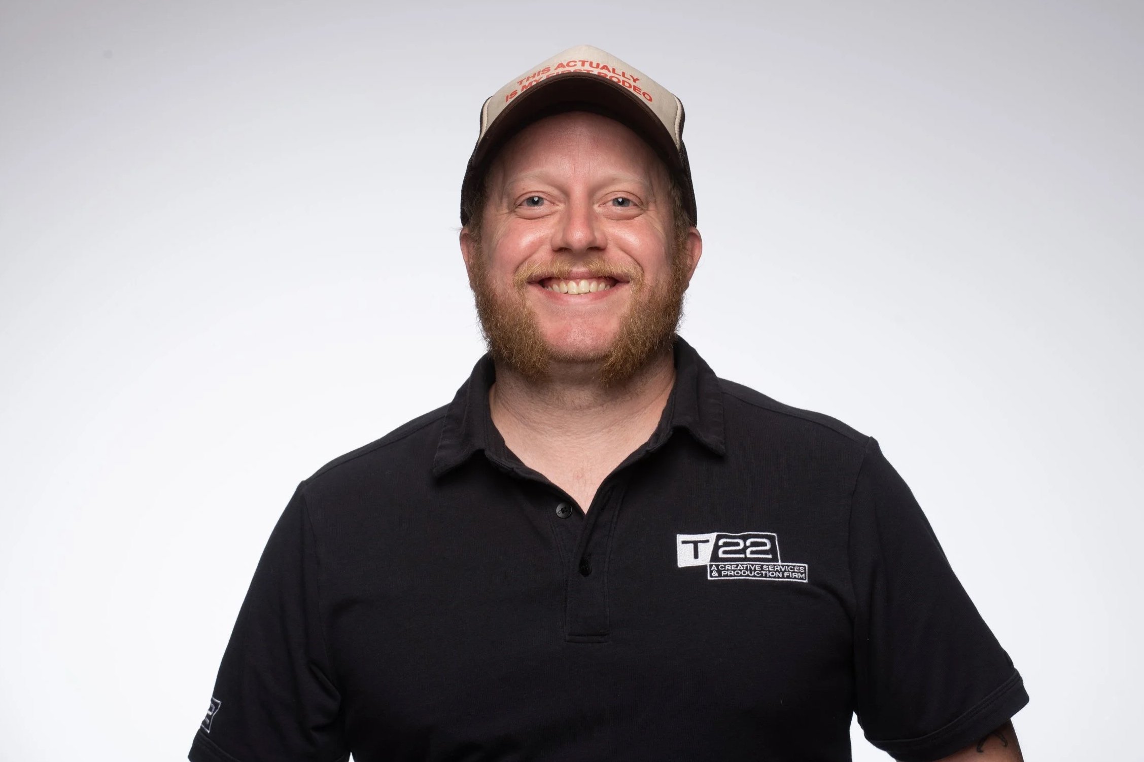 Man with a blonde red beard and a baseball cap wearing a black polo smiling at the camera.
