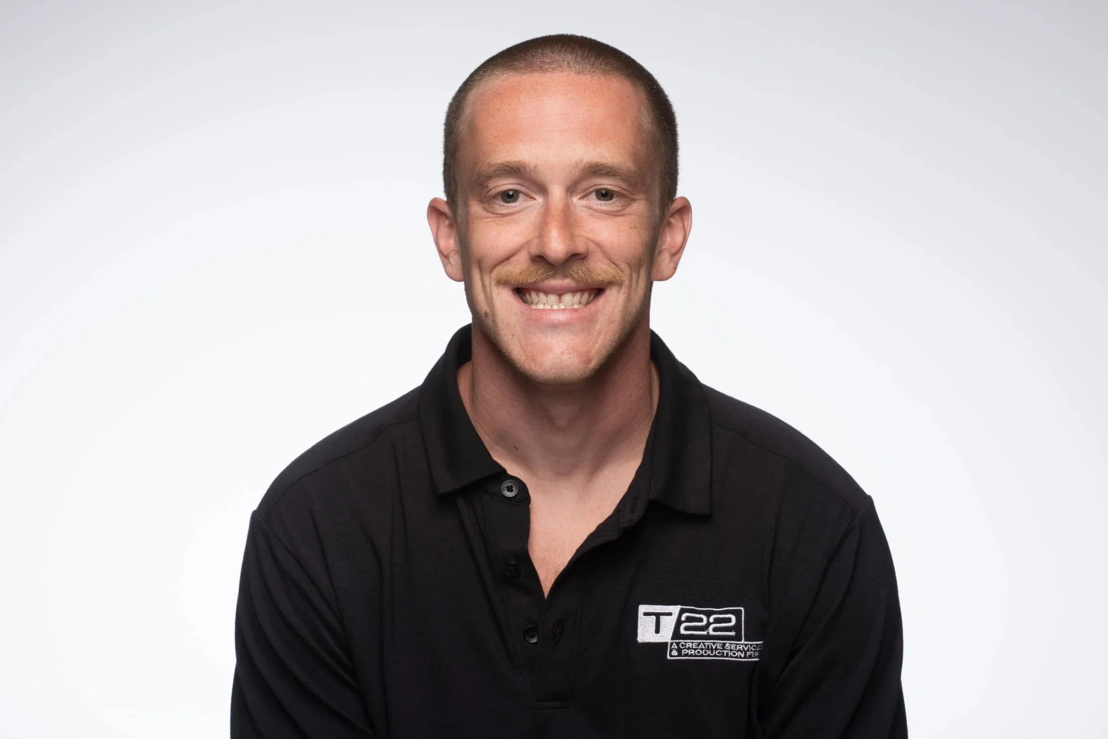 Young man with mustache smiling at camera wearing a T/22 polo who is an audio technician