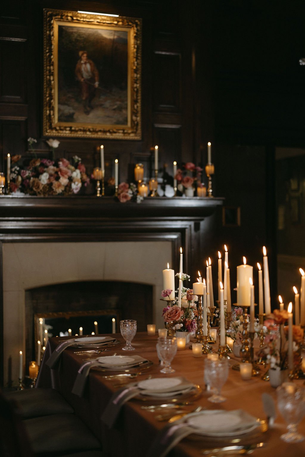 Antique stone fireplace mantel decorated with glowing taper candles, gold framed art, and warm floral touches.   