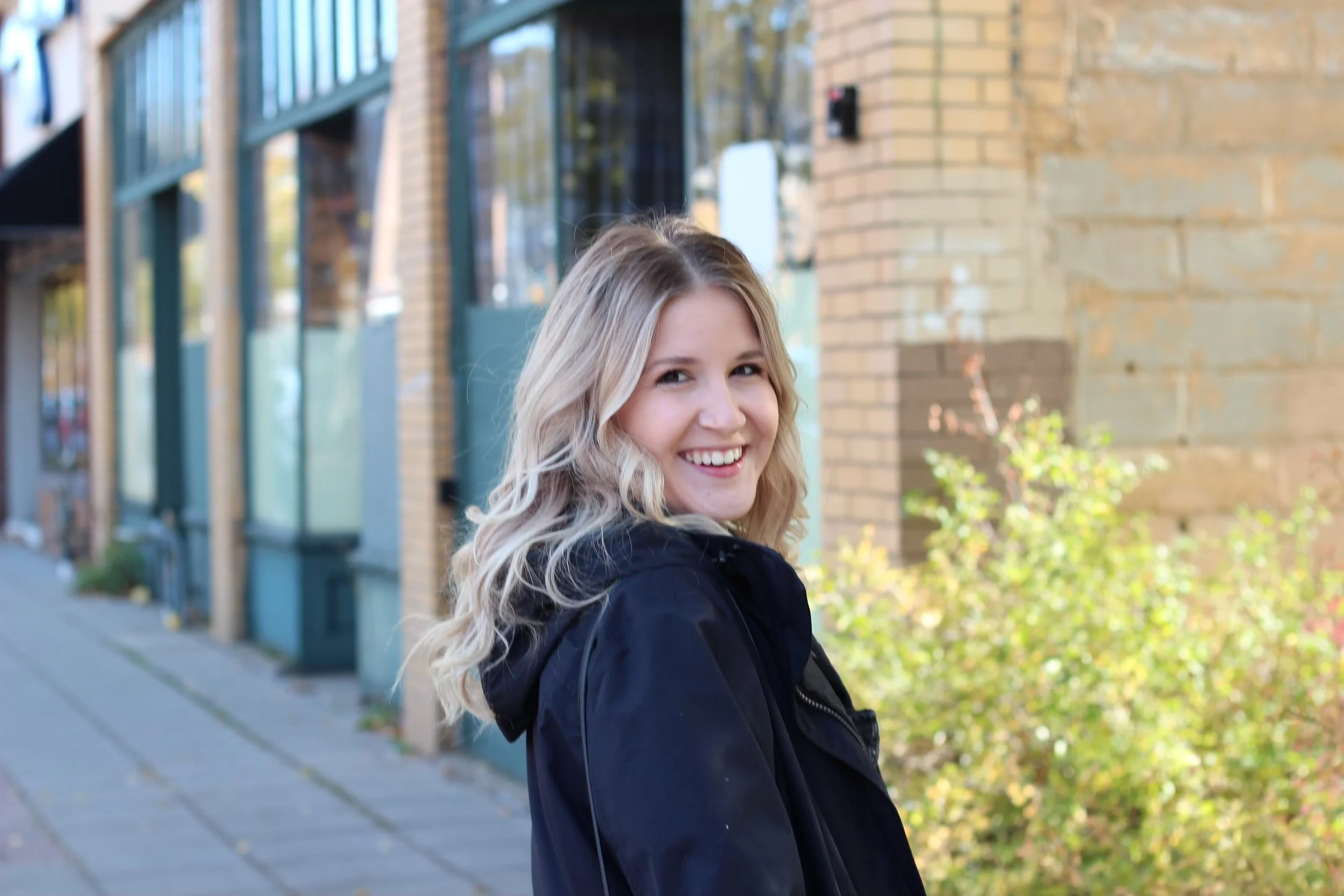 A woman with blonde curly hair smiling outdoors on a sidewalk, wearing a black jacket, with a brick building and greenery in the background.