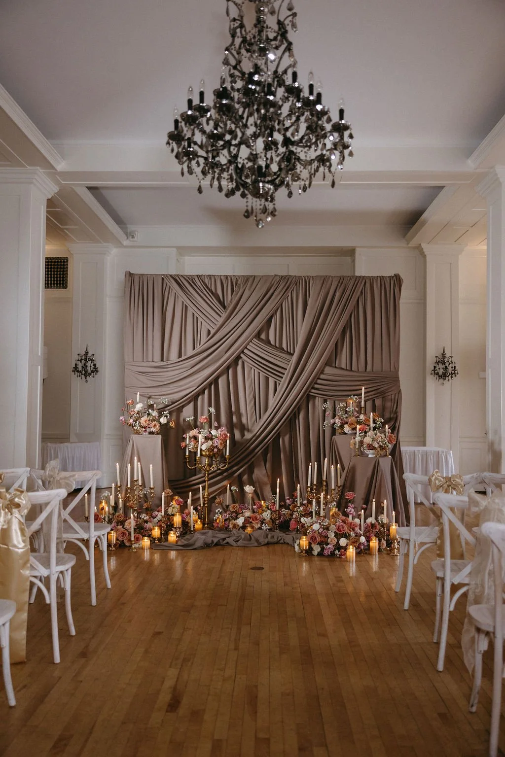 Moody vintage wedding ceremony venue styled with draped wall fabric, glowing candlelight along the aisle, and transparent acrylic ghost chairs.
