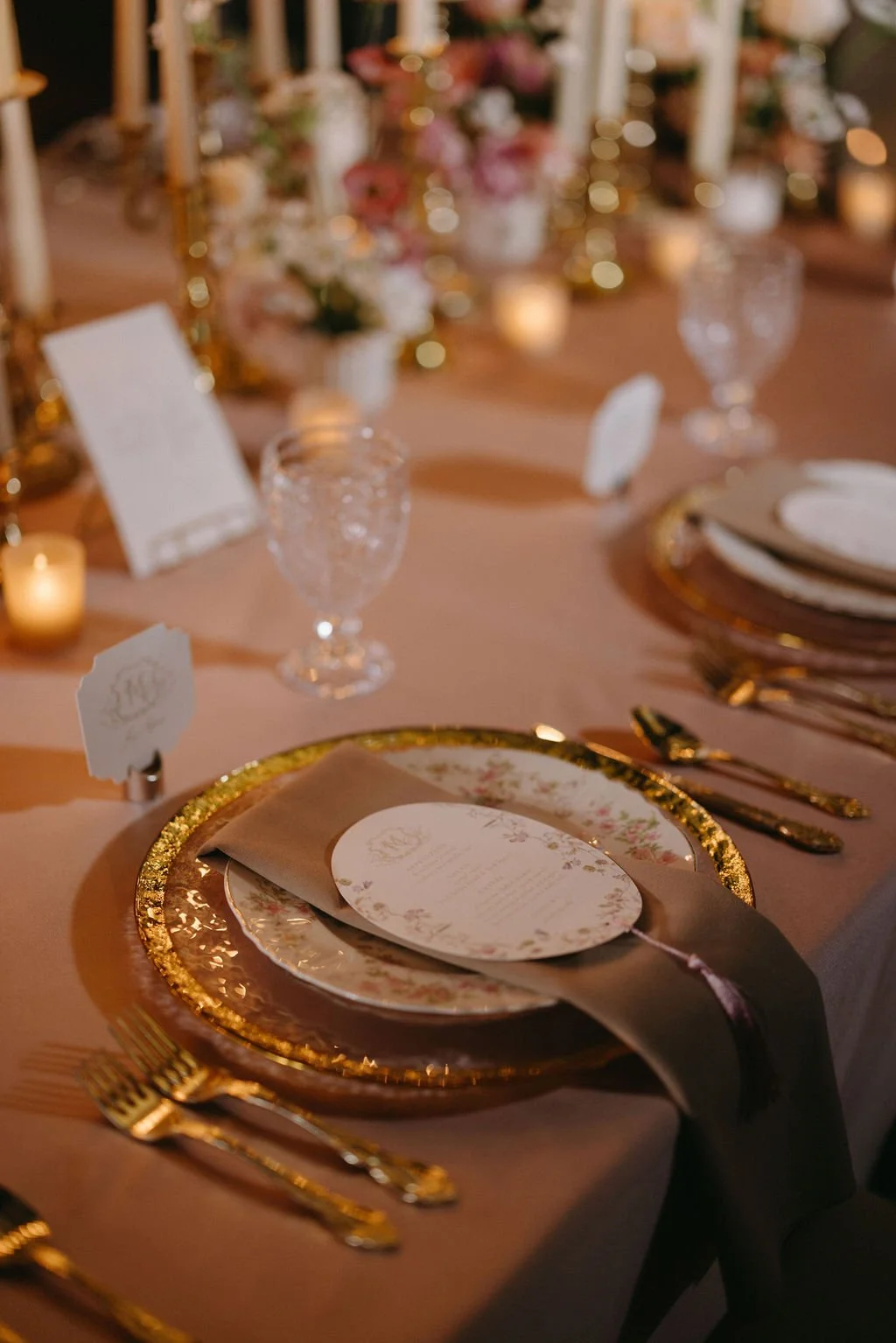Vintage wedding place setting featuring a gold charger plate, textured menu card, glass stemware, and warm candlelight.   