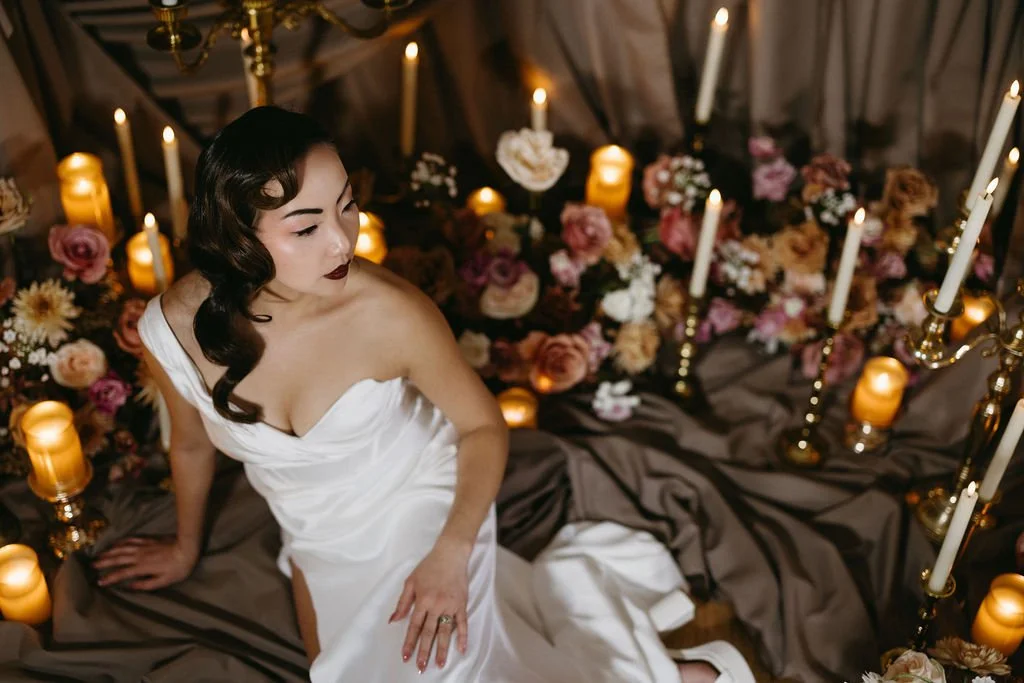 Vintage-inspired bride with finger wave hair styled in a strapless gown, reclining on draped brown fabric surrounded by glowing taper candles.