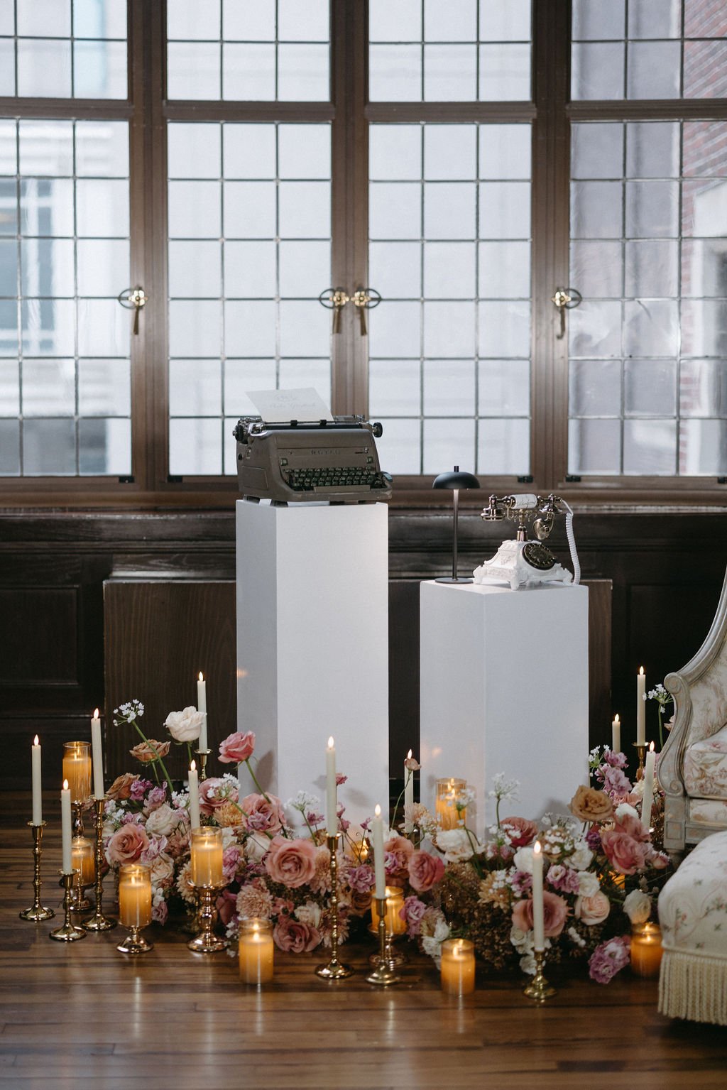 Vintage typewriter and rotary phone displayed on white pedestals, surrounded by floor candles and lush floral arrangements.   
