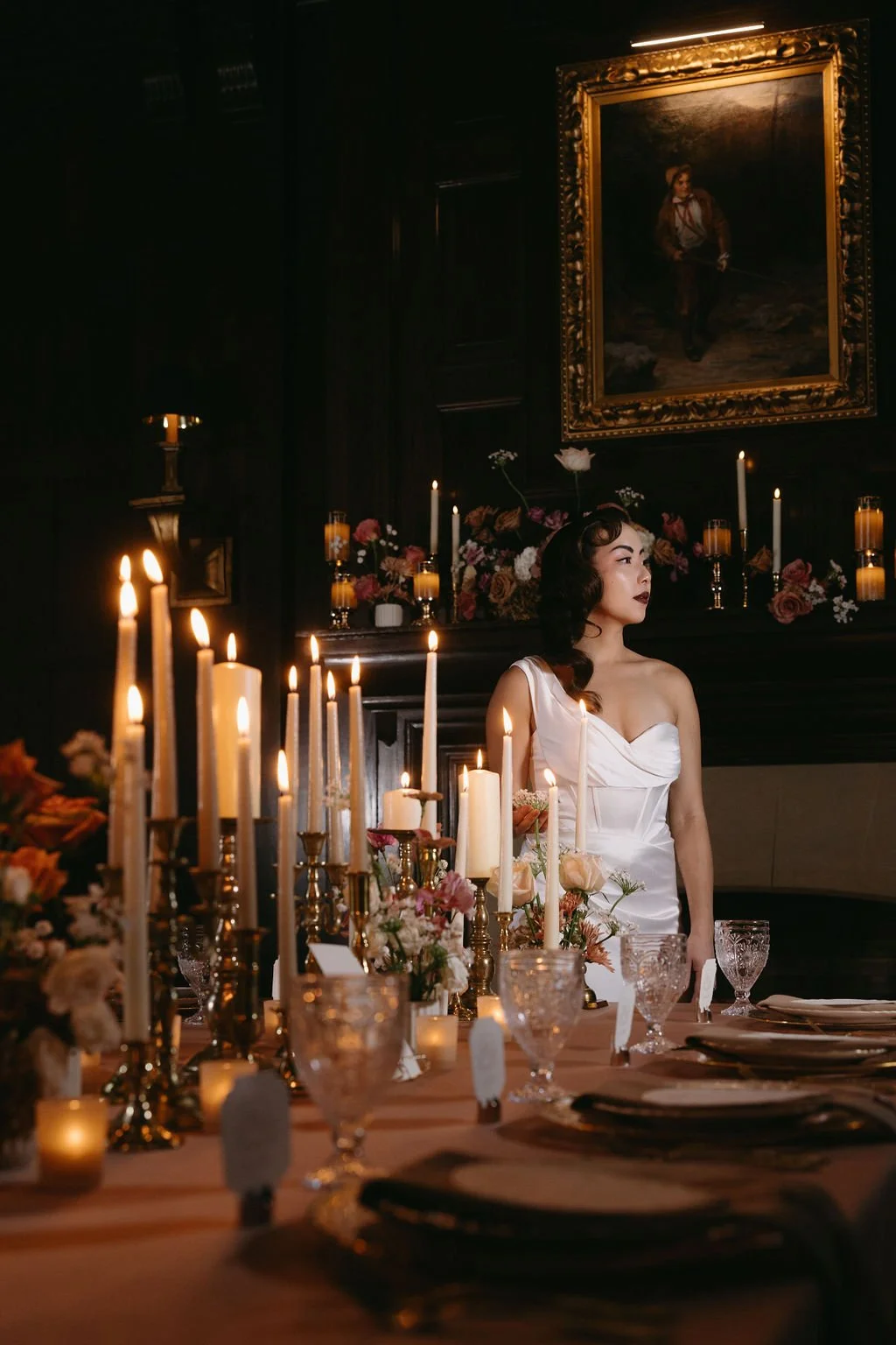 Bride in a silk wedding gown posing beside a floor-standing gold candelabra with flickering taper candles.