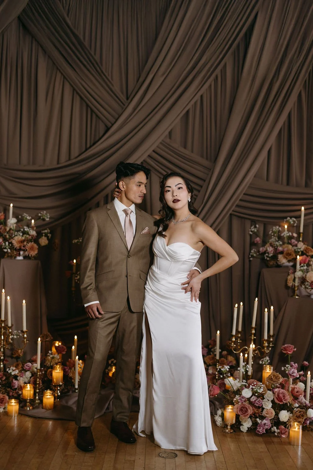 Bride and groom seated together against a draped fabric backdrop for a timeless vintage editorial portrait.