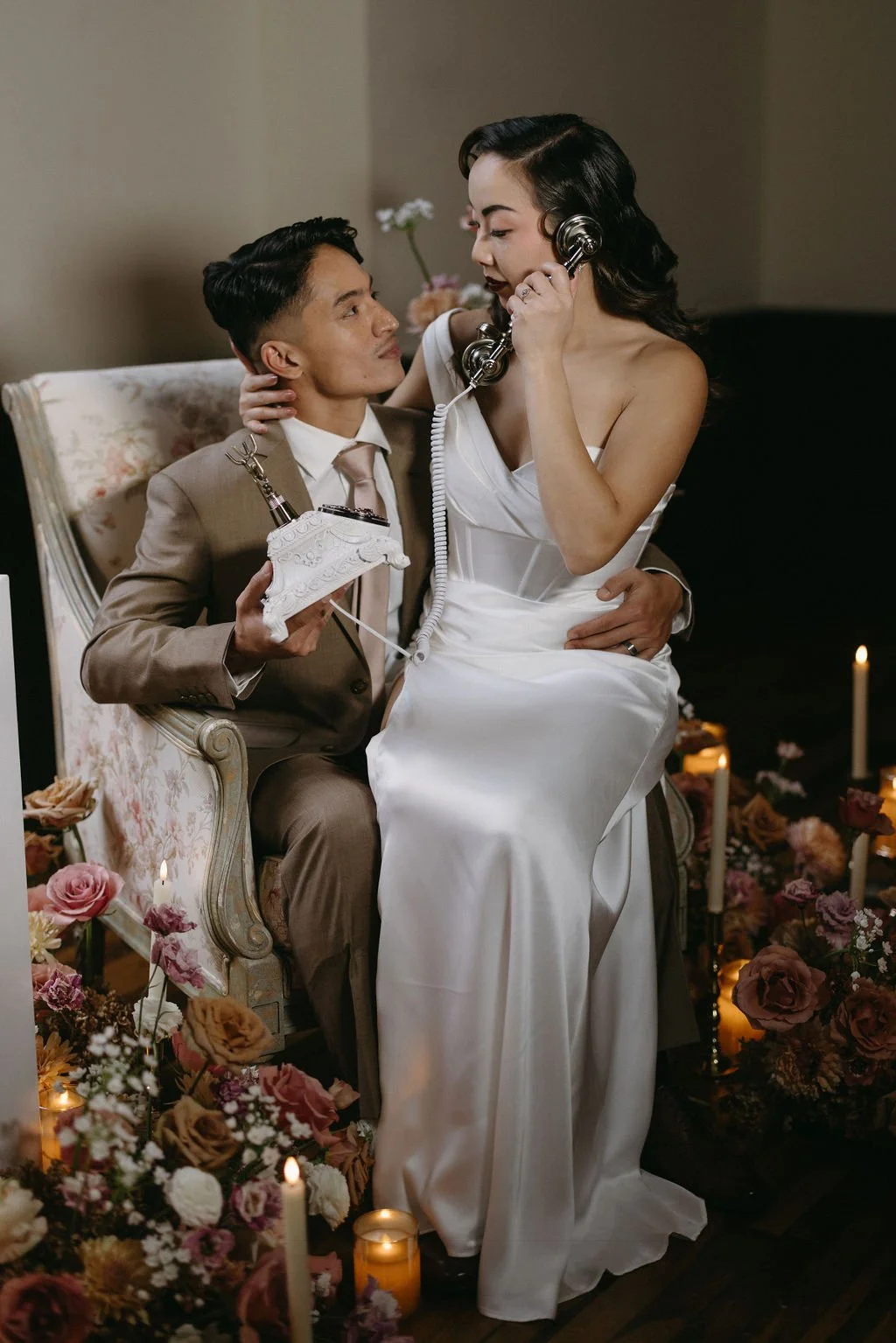 Playful vintage wedding portrait of the bride sitting on the groom's lap in an floral armchair while speaking on a white vintage rotary phone.