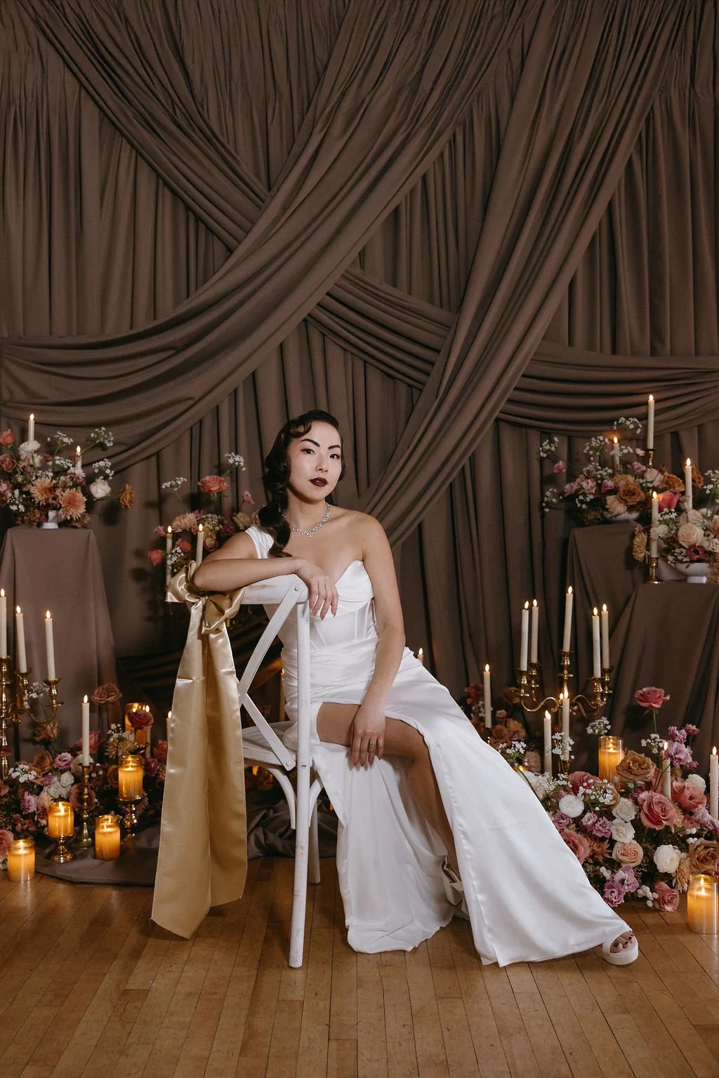 Vintage bride in a strapless gown sitting among glowing taper candles, draped backdrop fabrics, and warm floral arrangements by Revel & Flourish.